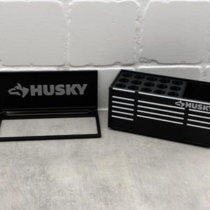 Può includere: Organizzatore di utensili nero con strisce bianche e il logo "HUSKY". L'organizzatore ha più fori circolari e un vano rettangolare. È presente anche una cornice nera separata con il logo "HUSKY".
