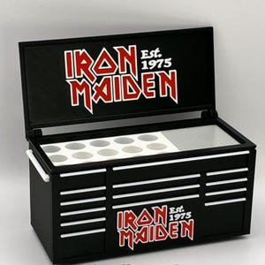 Può includere: Cassetta degli attrezzi nera con il logo Iron Maiden in rosso e bianco. La cassetta degli attrezzi ha un coperchio incernierato e più cassetti. È visibile anche il testo "Est. 1975". L'interno ha una sezione con supporti circolari.