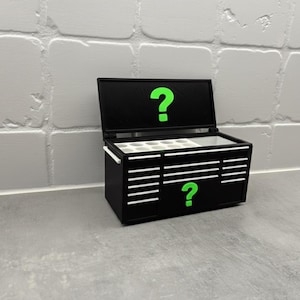 May include: Black toolbox with white drawer accents and a green question mark on the lid and front. The toolbox is open, revealing a white interior with compartments. The background is a white brick wall.