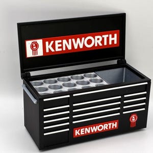 Può includere: Cassetta degli attrezzi nera con il logo Kenworth in rosso e bianco. La cassetta degli attrezzi ha un coperchio incernierato, un vano con supporti circolari e strisce bianche orizzontali. Il logo Kenworth è anche sulla parte anteriore.