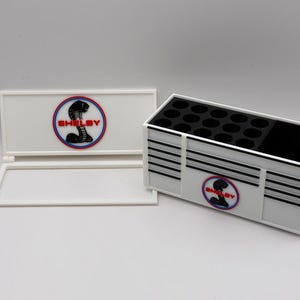 Può includere: Organizzatore rettangolare bianco con strisce orizzontali nere e bianche e un logo Shelby rosso, bianco e blu. L'organizzatore ha più fori circolari e un vano nero. È presente anche un coperchio abbinato.