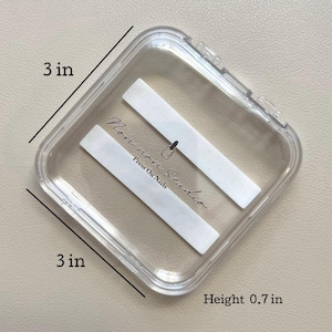 May include: A clear, square plastic case, 3 inches by 3 inches, with a height of 0.7 inches. Inside are two white rectangular inserts. The case has the text "Non non Studio Press On Nails" in black script.