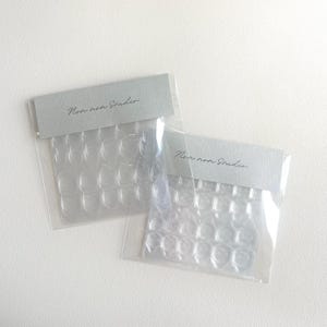 May include: Two clear plastic packages of press-on nails. Each package has a gray label with the text "Non non Studio" in a handwritten style. The nails are arranged in rows within the clear packaging.