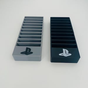 May include: Two console game storage organizers. One is gray with a black PlayStation symbol, the other is black with a white PlayStation symbol. Each organizer has multiple slots for game cases.