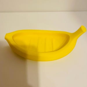 Pode incluir: Um saboneteira amarela brilhante em forma de banana. A saboneteira tem um design curvo com uma borda elevada e um pequeno caule em uma extremidade. O interior tem ranhuras verticais. A saboneteira é feita de material liso.