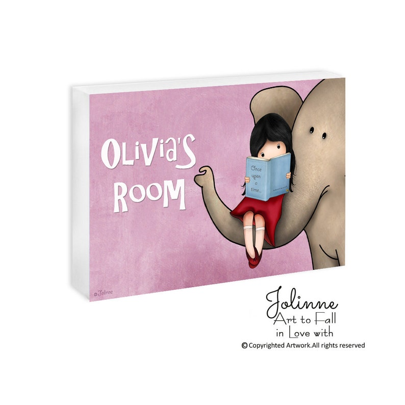 Op de afbeelding: Een roze en wit bord met de tekst "Olivia's Room" in wit. Een meisje met lang zwart haar zit op de rug van een olifant en leest een boek met de titel "Once upon a time..." De olifant is grijs met roze accenten. Het bord is versierd met een roze en wit bloemenpatroon.