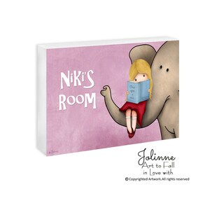Op de afbeelding: Een roze en wit gepersonaliseerd kamerschild met de tekst "Niki's Room" en een cartoonillustratie van een meisje dat op een olifant zit en een boek leest met de titel "Once Upon a Time".