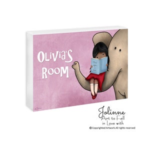 Op de afbeelding: Een roze en wit bord met de tekst "Olivia's Room" in witte letters. Een cartoonmeisje met bruin haar zit op een grijze olifant en leest een blauw boek met de tekst "Once upon a time..." Het bord is een schattige en fantasierijke toevoeging aan een kinderkamer.