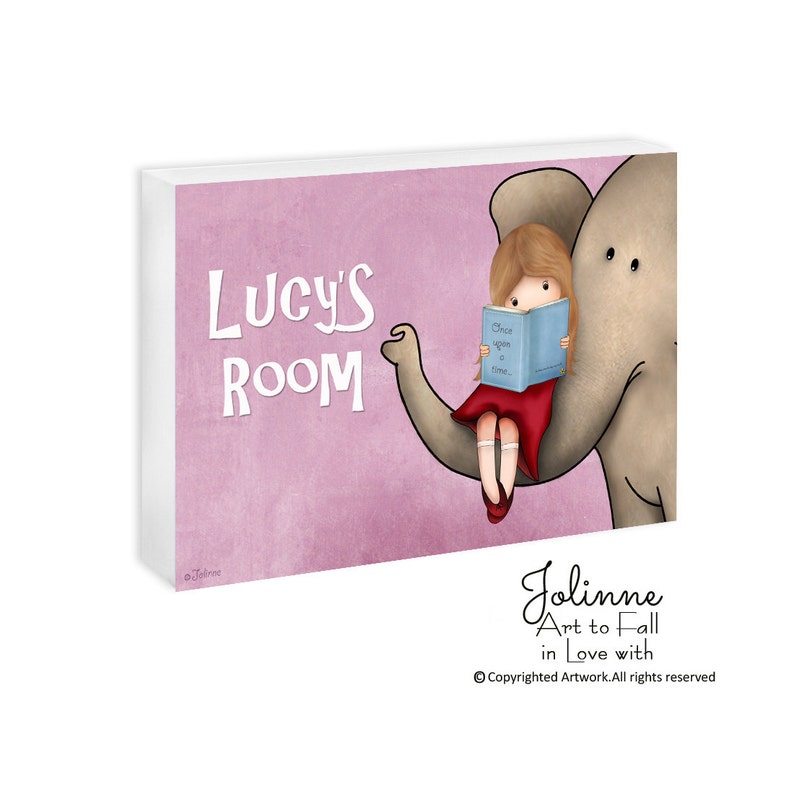 Op de afbeelding: Een roze en witte ge&iuml;llustreerde wandkunstprint met een meisje dat op de slurf van een olifant zit. Het meisje leest een boek met de tekst "Er was eens..." en de tekst op de print zegt "Lucy's kamer".