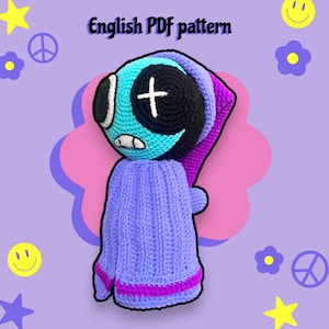 May include: A handmade crochet doll with a teal and black face, a white cross, and a purple hat and sweater. The doll has a whimsical design and the text "English PDF pattern" is at the top.