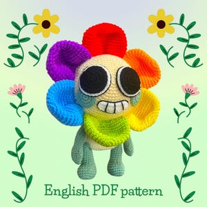 Könnte beinhalten: Ein gehäkeltes, blumenförmiges Plüschtier mit einem lächelnden Gesicht und Regenbogenblütenblättern. Das Spielzeug hat eine grüne Latzhose und Beine. Der Text "English PDF pattern" steht unten. Dekorative Blumen und Ranken befinden sich im Hintergrund.