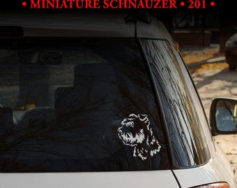 Peut inclure: Un autocollant en vinyle blanc représentant le profil de la tête d'un Schnauzer nain, appliqué sur la lunette arrière d'un véhicule blanc. Le texte MULTIPLE DESIGNS! MINIATURE SCHNAUZER 201 est affiché en blanc et rouge au-dessus de la voiture.