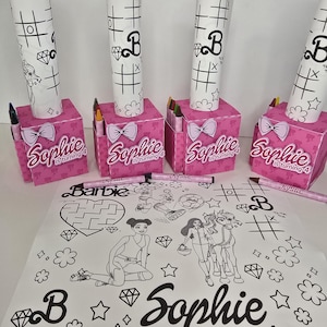 May include: Pink party favor boxes with white bow accents, each labeled "Sophie is turning 4." The boxes hold crayons and markers, topped with rolled-up coloring pages featuring tic-tac-toe and Barbie graphics. A large coloring sheet is also visible.