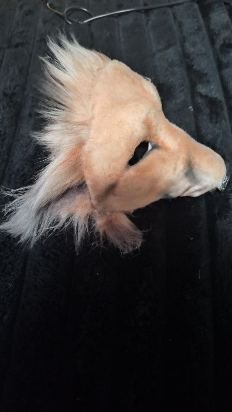 May include: A light brown, plush dog mask with a pointed snout and large eye openings. The mask has fluffy, light-colored fur around the ears and a darker shade on the nose. The mask is designed to be worn on the face.