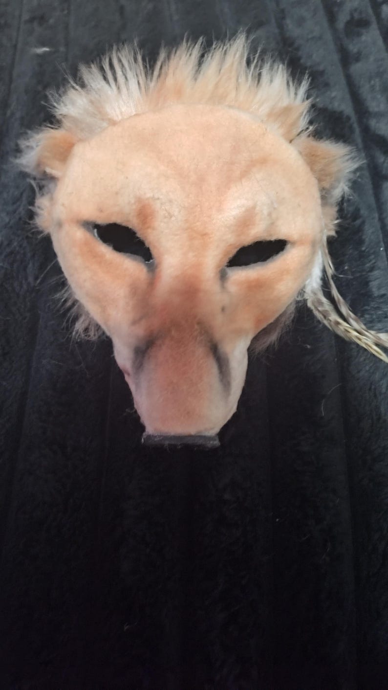 May include: A handmade mask in the shape of a lion's head. The mask is primarily tan with darker shading around the nose and eyes. It has a faux fur mane and feather accents.