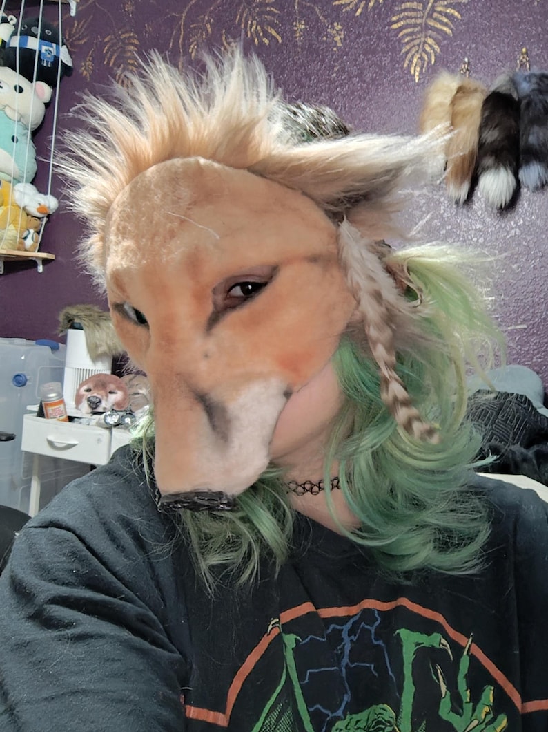 May include: A tan and brown fox mask with a faux fur mane and feather accents. The mask has a realistic design with detailed eyes and a black nose. The person wearing the mask has green hair and a black shirt.