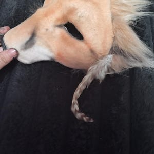 May include: A tan lion mask with a faux fur mane and tail. The mask has eye openings and is made of a soft, plush material. The tail is a mix of tan and brown. The mask is lying on a dark, textured surface.