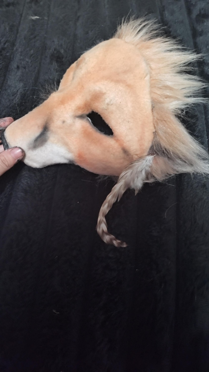 May include: A tan lion mask with a faux fur mane and tail. The mask has eye openings and is made of a soft, plush material. The tail is a mix of tan and brown. The mask is lying on a dark, textured surface.