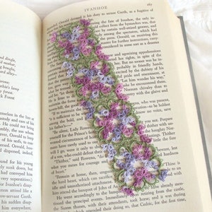 May include: A close-up of a book with a delicate lace bookmark. The bookmark is made of green and purple lace and features a floral design. The book is open to a page with text about the novel "Ivanhoe".