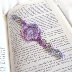 May include: A purple and green crocheted bookmark with a floral design. The bookmark is laying on a book with the text "Let him wander his way" visible.