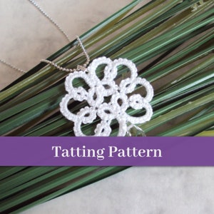May include: White tatting lace flower pendant on a silver chain. The pendant is hanging on a bed of green grass.