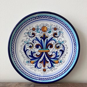 May include: A ceramic plate featuring a detailed floral design in blue, brown, and gold. The plate has a blue rim and a white centre with an ornate pattern. The plate is displayed on a wooden surface.