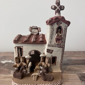 Puede incluir: Un belén de arcilla hecho a mano que representa el nacimiento de Jesús. La escena presenta una pequeña iglesia con una cruz en la parte superior, un pesebre con un bebé Jesús y varias figuras de personas y animales.