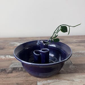 Puede incluir: Un jarrón de cerámica azul oscuro con tres aberturas para flores o vegetación. El jarrón tiene una base ancha y poco profunda y un cuello estrecho. Un solo tallo verde con hojas está en el jarrón.