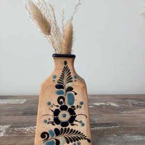 May include: A small, ceramic vase with a blue and green floral design. The vase is decorated with a brown glaze and has a rectangular shape. The vase is filled with dried pampas grass.