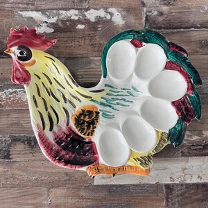 May include: A colourful ceramic egg holder in the shape of a rooster. The holder is painted with yellow, red, green, and black. It has a detailed rooster head and tail, with spaces for holding eggs. The design is decorative and functional.