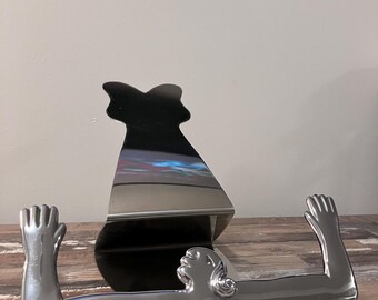May include: A silver metal bookend shaped like a woman with her arms raised. The bookend is reflective and has a smooth surface.