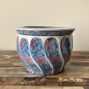 May include: A ceramic planter with a blue and white floral design. The planter features a repeating pattern of pink flowers and green leaves against a blue background. The rim has a blue geometric pattern.
