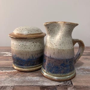 May include: A ceramic jug and a lidded jar. The pottery has a speckled, off-white glaze at the top, transitioning to tan, then a band of blue and brown. The jug has a handle and a flared spout. The jar is round.
