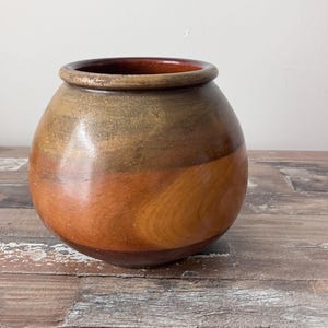 Puede incluir: Un jarrón redondo de madera con un esmalte marrón, naranja y beige. El jarrón está sobre una superficie de madera.