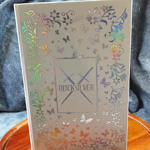 May include: A white journal with a holographic cover featuring a floral design with butterflies and the text "QUICKSILVER" in a silver font.