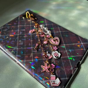 May include: A gold-toned phone charm with pink and white charms, including a heart, starfish, and bunny. The charm is attached to a phone case with a holographic plaid pattern. The charms are accented with pearls and pink enamel.