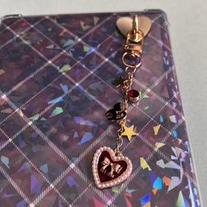 Pode incluir: Um pingente dourado com fecho em forma de coração e uma corrente pendurada. A corrente apresenta um gato preto, estrelas douradas, uma gema vermelha e um pingente em forma de coração com laço e borda perolada. O fundo é uma superfície holográfica com padrão xadrez.