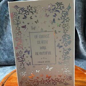 May include: A book with a light gray cover featuring a floral design with butterflies and the text "Of Course Death Was Beautiful" by Callie Hart.