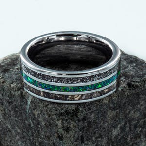 May include: A metallic ring featuring a central band of vibrant green crushed opal flanked by two textured dark gray inlay sections. The ring has a polished silver-toned finish on the edges and a smooth interior surface, creating a distinct contrast.