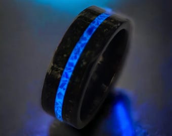 May include: A black ring featuring a textured, dark surface with a central, horizontal inlay that emits a bright, glowing blue light. The band has a smooth, polished interior and a dark, mottled exterior finish that contrasts with the vibrant blue glow.