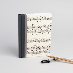 May include: A hardcover notebook featuring a cream-colored cover printed with black musical notation. The spine is bound in dark gray fabric. Two pens, one black and one metallic gold, are positioned in the foreground of the notebook.