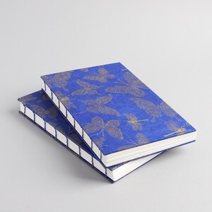 May include: Two blue notebooks with gold butterfly and dragonfly patterns. The covers have a textured appearance, and the pages are white. The notebooks are stacked on top of each other, with the spine visible.