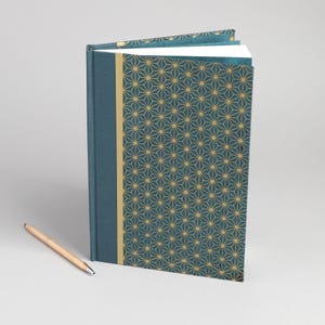 May include: A hardbound journal with a teal cover featuring a gold geometric star pattern and a solid teal spine with a gold vertical stripe, accompanied by a light wood-toned ballpoint pen positioned diagonally on the surface.