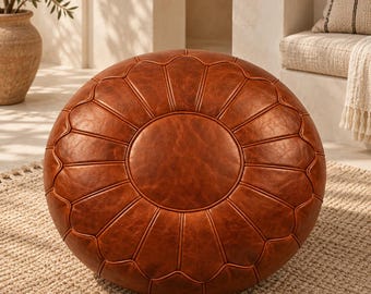 Può includere: Un pouf rotondo in pelle color marrone cognac caratterizzato da un pannello centrale circolare circondato da segmenti radianti a forma di petalo. La superficie presenta una finitura testurizzata con cuciture ben visibili.