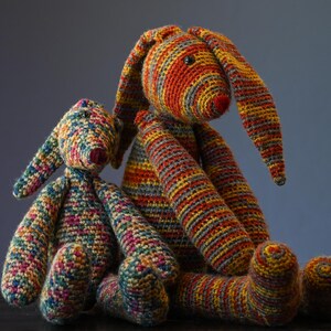May include: Two handmade crochet bunny rabbits in vibrant colors. The larger bunny is primarily orange, yellow, and gray, while the smaller one features a mix of blues, greens, and pinks. Both have long ears and red noses.