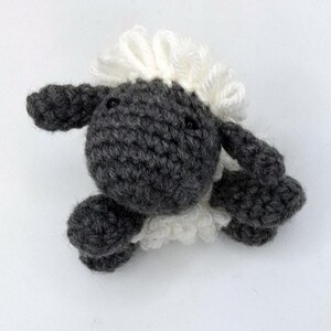 Op de afbeelding: Een handgemaakt gehaakt schaap speelgoed. Het schaap heeft een grijze lichaam, hoofd en ledematen, met een pluizige witte wollen manen en staart. Het schaap heeft kleine zwarte ogen en ligt. Het speelgoed is gemaakt van zacht garen.