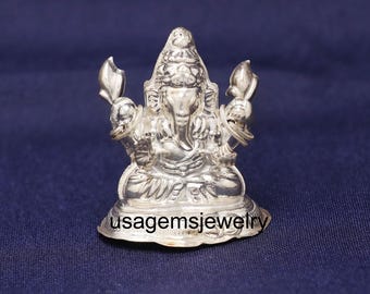 Può includere: Una dettagliata statuetta d'argento della divinità indù Ganesha, seduta con più braccia e una testa di elefante. La statuetta presenta dettagli intricati ed è ambientata su uno sfondo blu scuro. La base della statuetta riporta la scritta "usagemsjewelry".