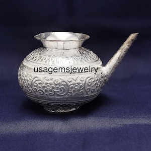 May include: A silver-colored, ornate water pot with a spout, set against a dark blue background. The pot features detailed floral patterns and the text "usagemsjewelry". The vessel has a rounded body and a flared rim.