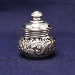 Può includere: Un piccolo barattolo color argento con coperchio. Il barattolo ha un corpo arrotondato con intagli floreali ornati e il testo "usagemsjewelry". Il coperchio è a cupola con un piccolo pomello in cima. Lo sfondo è blu scuro.