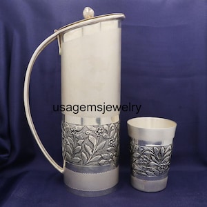 Puede incluir: Un juego de jarra y taza de plata. La jarra tiene un asa curva y una tapa con un pequeño pomo. Ambas piezas presentan un diseño floral alrededor de la base. El texto "usagemsjewelry" es visible.